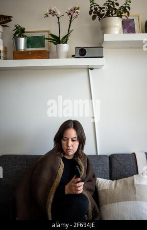 Frau eingewickelt in Decke sitzend auf der Couch, die weit entfernt aussach Stockfoto