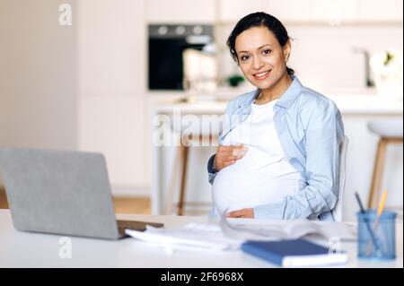 Schwanger Geschäftsfrau. Glücklich selbstbewusst Attraktive Mixed Race Schwangere, Managerin, Immobilienmaklerin, Designerin, die aus der Ferne arbeitet, sitzt am Arbeitsplatz, hält den Bauch mit den Händen und schaut in die Kamera Stockfoto