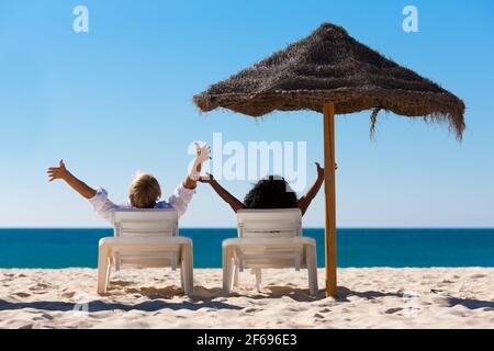 Pärchen sitzt in Liegestühlen unter einem Sonnenschirm am Strand, der sich die Arme streckt und sich frei fühlt Stockfoto