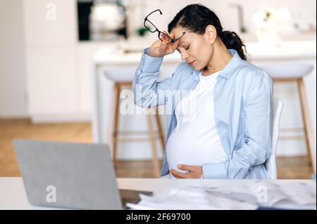 Müdigkeit, stressige Mischtrassen reife Schwangere, freiberufliche Mitarbeiterin, Designerin oder Managerin, die aus der Ferne arbeitet, sitzt am Schreibtisch, macht Pause, hat Kopfschmerzen, hält Brille in der Hand, die Augen geschlossen Stockfoto