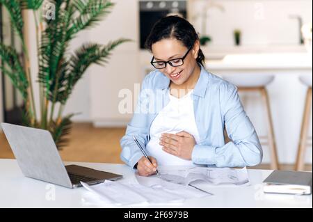 Selbstbewusste, schöne, glückliche Schwangere mit Brille, gemischter Rasse, Freiberuflerin, Managerin oder Designerin, die aus der Ferne arbeitet, Verwendet Laptop, Notizen machen und überprüfen Design-Projekt, streicheln Bauch, lächeln Stockfoto