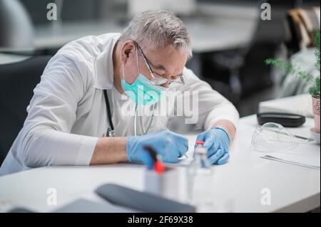 Ein erfahrener Arzt für Infektionskrankheiten füllt Dokumente an seinem Schreibtisch aus, in einer Privatklinik während der Arbeit. Stockfoto