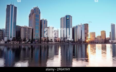 Panaromischer Blick auf die wolkenkratzer von sharjah, die wunderschöne Spiegelungen im Wasser zeigen, die während des frühen Morgensonnenaufgangs von der Insel Al Noor aufgenommen wurden Stockfoto