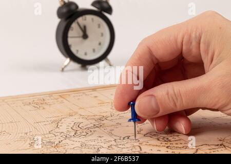 Weibliche Hand zeigt mit einer Nadel den Ort der Reise auf der Weltkarte. Auf einem weißen Holztisch ein schwarzer Wecker. Nahaufnahme. Selektiver Fokus Stockfoto