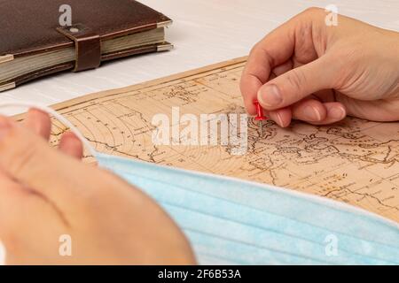 Weibliche Hand zeigt mit einer Nadel den Ort der Reise auf der Weltkarte. Auf einem weißen Holztisch blaue medizinische Maske und Tagebuch. Nahaufnahme. Selektiver Fokus Stockfoto