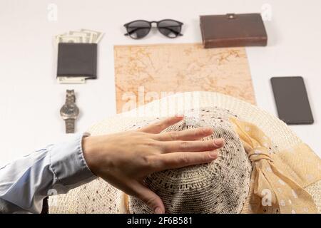 Weibliche Hand hält einen Strohhut. Auf einem weißen Holztisch liegt eine Weltkarte, eine schwarze Geldbörse mit Geld, ein Tagebuch, eine Uhr, ein Smartphone und eine Sonnenbrille. sel Stockfoto