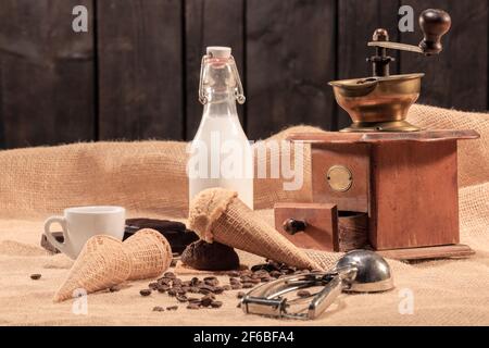 Verführerischer Eiskonus mit Kaffeebohnen, einer Flasche Milch, einer Kaffeetasse und einer klassischen Kaffeemühle. Selektiver Fokus. Eis- und Süßwarenkonzept. Stockfoto