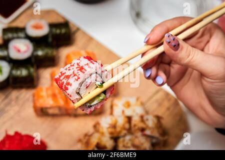 Mann, der Essstäbchen-Sushi-Rolle auf dem Hintergrund einer Reihe verschiedener Sushi-Rollen hält. Nahaufnahme, selektiver Fokus Stockfoto