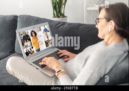 Seitenansicht eine Unternehmerin mittleren Alters mit Laptop für Videoanruf in entspannter Atmosphäre auf dem Sofa zu Hause liegend, ältere Frau in Videokonferenz beteiligt, beobachten Bildungskurse zu Hause Stockfoto