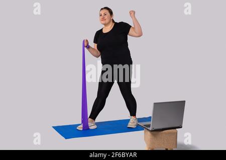 Fitness mit Gummiband. Eine fette Frau in Sportbekleidung trainiert mit einem Gummiband auf der Yogamatte gegenüber dem Laptop. Gesunder Lebensstil. Sport und Stockfoto