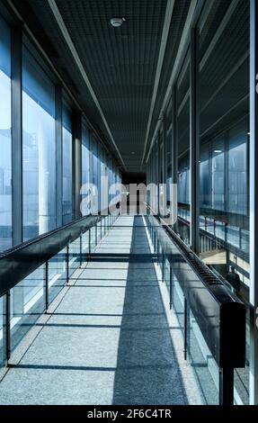 Langer, enger Korridor, der außerhalb des hohen Gebäudes führt. Die Glaswand bietet einen perfekten Blick auf die Umgebung und bringt natürliches Licht in die Büros. Stockfoto