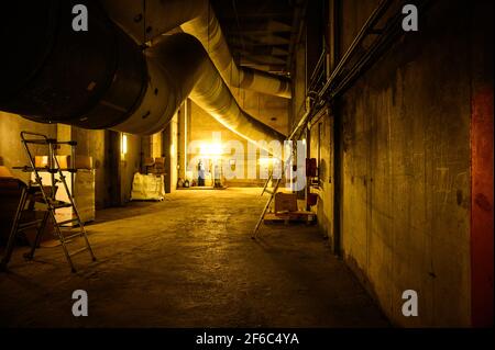 Industriehintergrundfoto bei schwachem Licht. Versorgungskorridor mit massiven Rohren des Belüftungssystems, beleuchtet durch gelbes Licht.Korridor Stockfoto