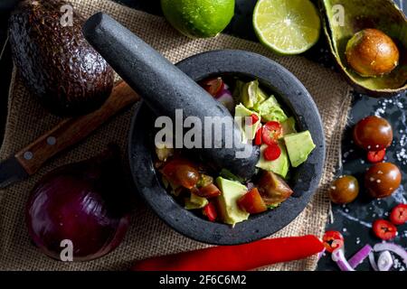 Zubereitung von Guacamole in einem traditionellen Steinmörtel mit allen Zutaten (gehackte Avocados, Limette, Zwiebel, Tomaten und Chilischoten). Appetizin Stockfoto