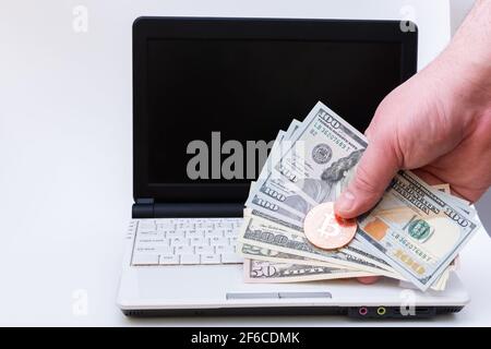 Die Hand hält Bitcoin- und 100-Dollar-Papierrechnungen über dem Laptop mit schwarzem Bildschirm auf weißem Hintergrund. Bitcoin Kryptowährung Konzept. USD in BTC. V Stockfoto