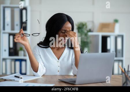 Schwarze junge Geschäftsfrau mit müde gereizten Augen, sitzen am Schreibtisch mit Laptop, erschöpft von Online-Arbeit im Büro Stockfoto