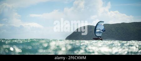 Ein Mann Flügel Folien auf dem Meer mit einem Hand gehalten aufblasbaren Flügel, Reiten ein Tragflächenboot Surfbrett. Türkisfarbenes Meer, wolkig blauer Himmel. Stockfoto