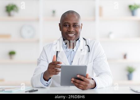 Online-Beratung. Fröhlicher schwarzer Arzt mit digitalen Tablet in seinem Büro, im Gespräch mit Patienten entfernt während der Quarantäne. Stockfoto