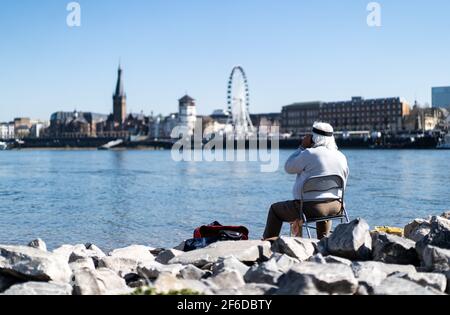 Düsseldorf, Deutschland. März 2021, 31st. Ein Angler sitzt am Rheinufer in Oberkassel, während das Riesenrad am Burgplatz im Hintergrund zu sehen ist. Meteorologen prognostizieren frühlingshaftes Wetter für heute mit Temperaturen bis zu 25 Grad. Quelle: Jonas Güttler/dpa/Alamy Live News Stockfoto