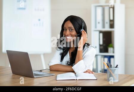 Lächelnde Geschäftsfrau mit Headset, die auf die Kamera schaut und den Laptop für die Online-Kommunikation im Büro nutzt Stockfoto
