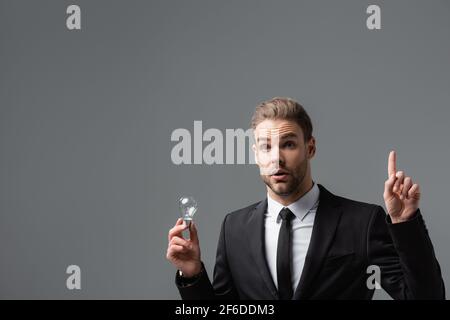 Aufgeregter Geschäftsmann, der eine Idee zeigt, während er die Glühbirne isoliert hält Auf grau Stockfoto