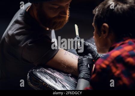 Ein Tattoo-Künstler mit kurzen Haaren und Piercings bereitet sich darauf vor, ein Tattoo auf den Arm eines Mannes in einem Tattoo-Salon zu bekommen. Stockfoto
