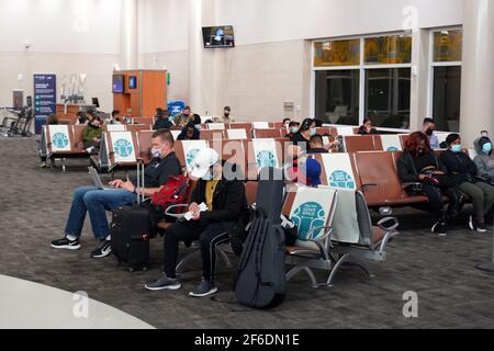 Menschen mit Gesichtsmasken sitzen zwischen physischen und sozialen Distanzierungszeichen mit den Worten „Stay Safe“. Platz lassen“ auf den Sitzen in Terminal B des Stockfoto