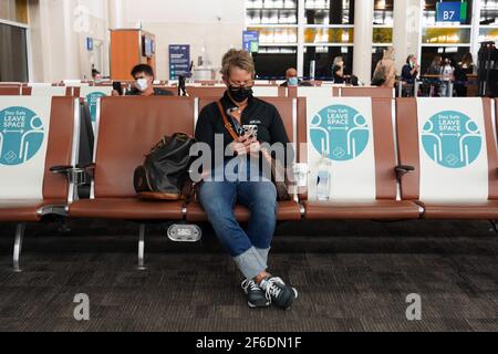 Menschen mit Gesichtsmasken sitzen zwischen physischen und sozialen Distanzierungszeichen mit den Worten „Stay Safe“. Platz lassen“ auf den Sitzen in Terminal B des Stockfoto