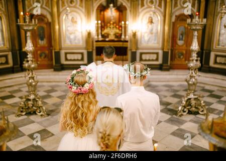 Der Priester, der Bräutigam und die Braut in Kränzen stehen während der Hochzeit auf dem Altar der St. Nikolaus-Kirche in Kotor, Rückansicht Stockfoto