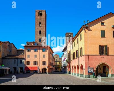 Blick auf den gepflasterten Stadtplatz zwischen alten bunten Häusern und mittelalterlichen Türmen unter blauem Himmel in Alba, Piemont, Norditalien. Stockfoto