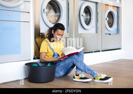 Eine lateinische Frau las ein Buch und saß auf dem Boden, während sie auf die Wäsche in der Selbstbedienungswäsche wartete. Sie lächelt. Stockfoto