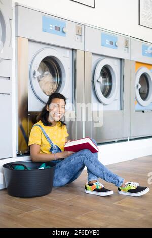 Junge lateinamerikanerin wartet darauf, dass ihre Kleidung im Waschsalon zur Selbstbedienung gewaschen wird. Sie lächelt und hat ein Buch in ihren Händen. Stockfoto