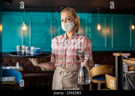 Kaffee und Wasser während des Corona-Virus servieren. Porträt einer Kellnerin Frau mit einem Pastellhemd und Schürze mit einer schützenden Gesichtsmaske zu Fuß aroun Stockfoto