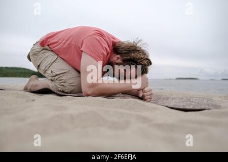 Der ruhige Mann macht Yoga, er macht die Übung, auf dem Teppich, am sandigen Ufer des Sees. Gesundes Lifestyle-Konzept. Stockfoto