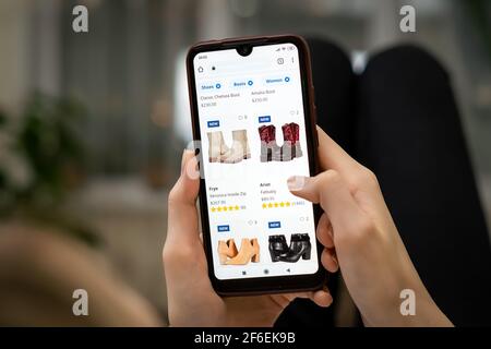 Frau liegt auf dem Sofa im Wohnzimmer und wählt ihre Schuhe über eine Online-Handy-App aus. Online-Shopping-Konzept Stockfoto