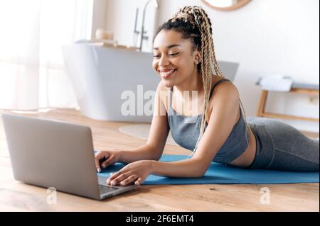 Athletisch schöne junge afroamerikanische Frau mit einer guten Figur, liegt zu Hause auf dem Boden, sieht Online-Fitnesskurse mit Laptop, geht in den Sport, führt einen gesunden Lebensstil, lächeln Stockfoto