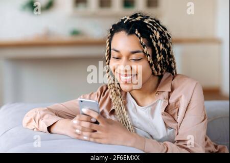 Glückliches, schönes afroamerikanisches Mädchen, das auf dem Sofa sitzt, in legerer Kleidung, Smartphone benutzt, Zeit in sozialen Netzwerken verbringt, lustige Videos anschaut, mit Freunden oder Freund chattet, lächelt Stockfoto