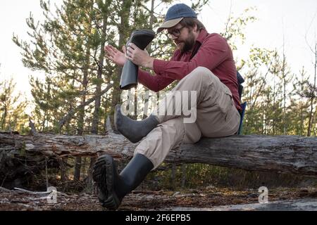 Der Mann in Wanderkleidung saß auf einem alten Baumstamm im Wald, zog seine Gummistiefel aus und schüttelte Müll aus. Aktives Lifestyle-Konzept. Stockfoto