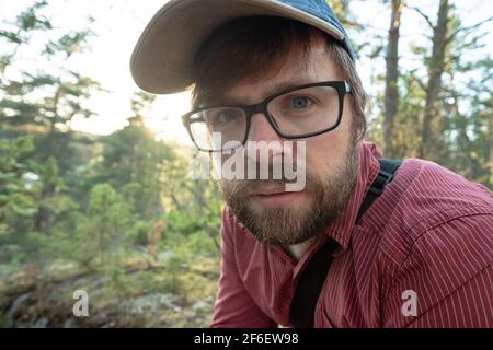 Bärtiger Mann mit Brille und Mütze, der nach einem langen Spaziergang im Wald ruht. Er schaut aufmerksam und nachdenklich. Nahaufnahme. Stockfoto