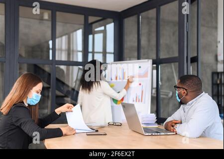 Kollegen tragen medizinische Masken während des Arbeitstages im Büro. Multirassisches Team, das an einem neuen Projekt im modernen Coworking Space arbeitet und sich mit Masken vor Viren schützt Stockfoto