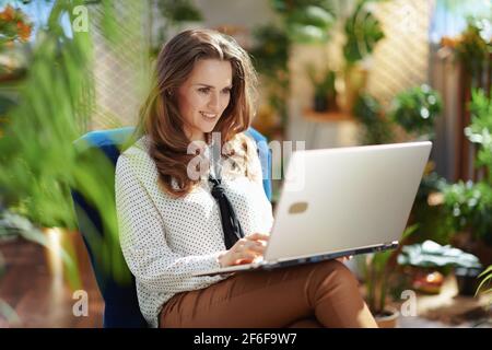 Green Home. Lächelnd stilvolle Mittelalterhausfrau mit langen welligen Haaren mit Laptop in beige Hosen und Bluse in modernen Hause in sonnigen Tag. Stockfoto