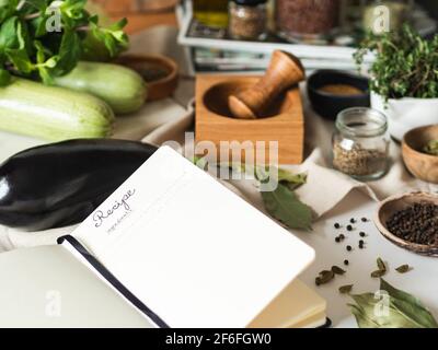 Offenes Notizbuch für Kochrezepte und verschiedene vegetarische Zutaten auf dem Küchentisch. Vorderansicht Stockfoto