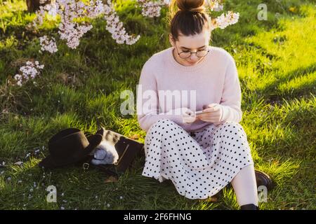 Junge kaukasische Hipster Mädchen in Brille und leichte Kleidung auf grünem Gras in der Nähe blühenden Baum im Park sitzen und mit ihrem Smartphone. Frau schreibt Stockfoto