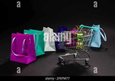 Kleiner Metalleinkaufwagen voller Geschenke, bunte Taschen, isoliert im Dunkeln, Online-Shopping, Winterverkauf, Supermarkt, Rabatt-Promotion und Black Frid Stockfoto