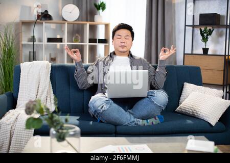 Entspannter asiatischer Mann, der auf der Couch sitzt, mit einem Laptop auf den Knien und mit geschlossenen Augen meditiert. Müde Freiberufler, die Pause während des Arbeitsprozesses zu Hause. Stockfoto