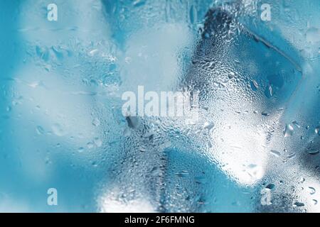 Eiswürfel in einem vernebelten Glas mit Tropfen Eiswasser Nahaufnahme Makro. Erfrischendes und kühlendes Getränk bei heißem Wetter. Blauer Hintergrund. Vollbild Stockfoto