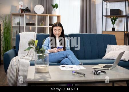 Attraktive asiatische Frau sitzt auf der Couch und mit modernen Smartphone. Freiberufliche Mitarbeiterin, die während des Arbeitsprozesses Pause einnahm. Tisch mit Dokumenten und Laptop in der Nähe. Stockfoto
