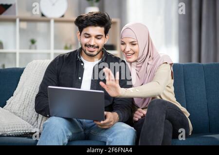 Lächelndes, glückliches muslimisches Paar mit Laptop-Computer zu Hause, winkend die Hände, während sie einen Videoanruf mit ihren Freunden oder ihrer Familie führen. Bleib zu Hause während einer Pandemie. Stockfoto