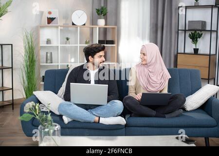 Zielstrebig junge lächelnde, glückliche muslimische Paare oder Freunde, die mit Laptops auf einer weichen Couch zu Hause in Lotusposition sitzen und sich gegenseitig schauen, während sie gemeinsame Freizeit verbringen, arbeiten oder online studieren. Stockfoto