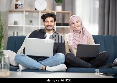 Ein junges, lächelndes, multiethnisches Paar im Alter von 30 Jahren, ein bärtiger Inder und eine muslimische Frau in Hijab, die in Lotuspose mit Laptops auf einer bequemen Couch zu Hause sitzen und in die Kamera blicken. Stockfoto