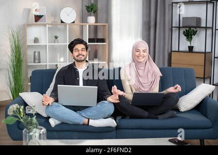 Sympathischer junger arabischer muslim in Hijab, sitzend in Lotuspose mit Laptop-pc auf dem Sofa im Wohnzimmer, meditierend mit geschlossenen Augen und Lächeln, Finger in Mudra faltend. Stockfoto